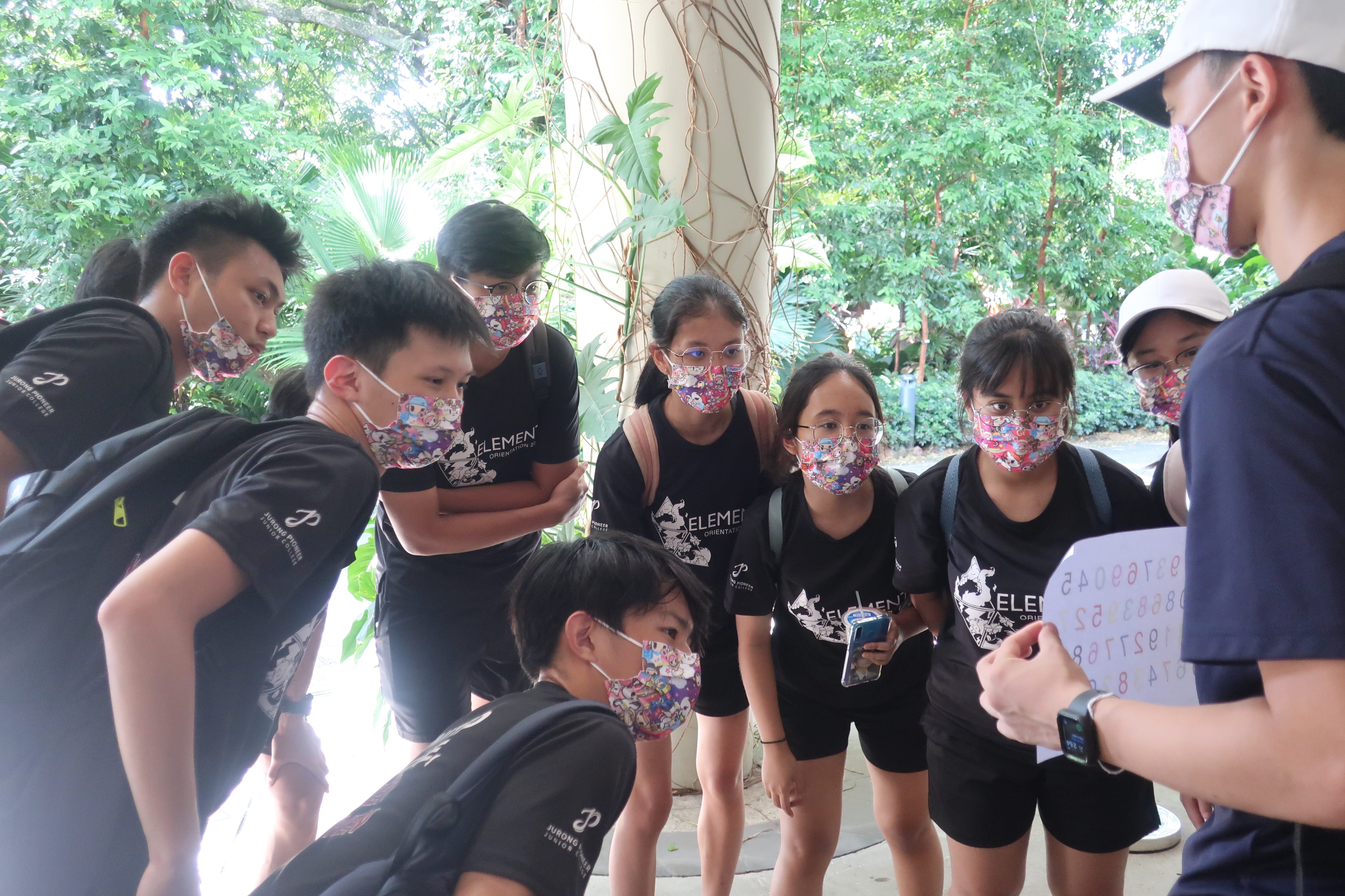 Masked teens in black shirts gather around a man holding a sheet of numbers outdoors by greenery.